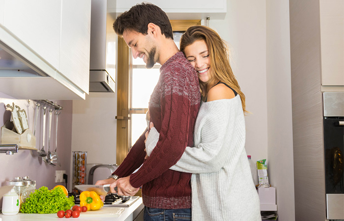 A smiling woman hugging her partner from behind while her partner is cutting vegetables.